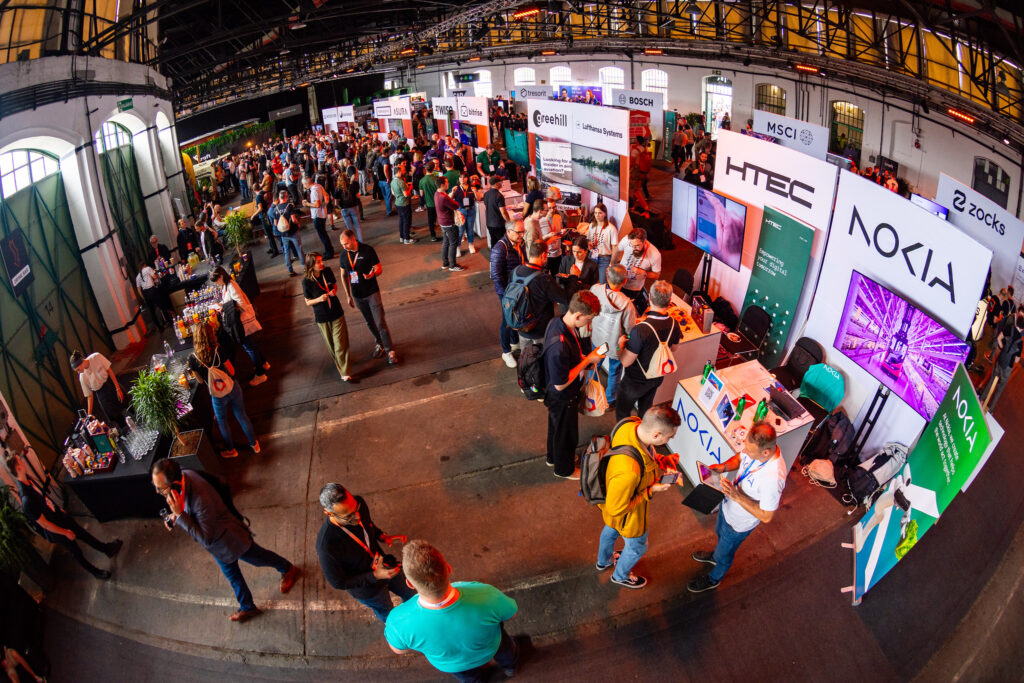 A vibrant exhibition area at Craft Conference with attendees networking, exploring sponsor booths like Nokia and HTEC, and enjoying refreshments. The industrial-style venue has high ceilings, large green doors, and bright lighting, creating a lively and engaging atmosphere.
